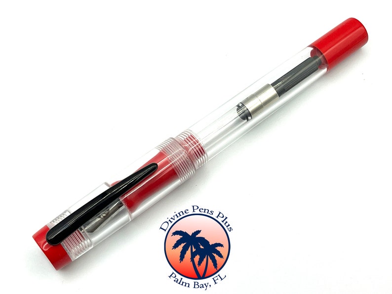 May include: A clear fountain pen with red accents. The pen has a transparent barrel, a black nib, and a red cap and end. The pen has a clip and a silver-colored metal section. The pen is a writing instrument.