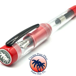 May include: A clear fountain pen with red accents and a black clip. The pen has a transparent barrel, allowing a view of the internal components. The pen's cap and end are red, with the brand name and logo visible on the end.