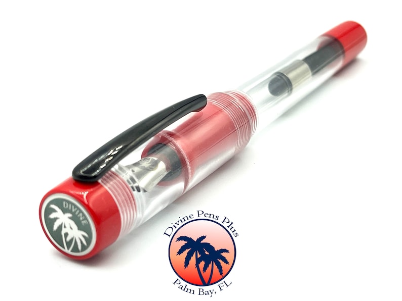 May include: A clear fountain pen with red accents and a black clip. The pen has a transparent barrel, allowing a view of the internal components. The pen's cap and end are red, with the brand name and logo visible on the end.