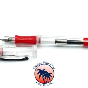 May include: A clear fountain pen with red accents, including the grip and cap. The pen's body is transparent, revealing the internal ink mechanism. The nib is silver-colored, and the cap is detached, lying next to the pen. The pen has the Divine Pens Plus logo.