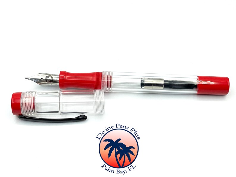 May include: A clear fountain pen with red accents, including the grip and cap. The pen's body is transparent, revealing the internal ink mechanism. The nib is silver-colored, and the cap is detached, lying next to the pen. The pen has the Divine Pens Plus logo.