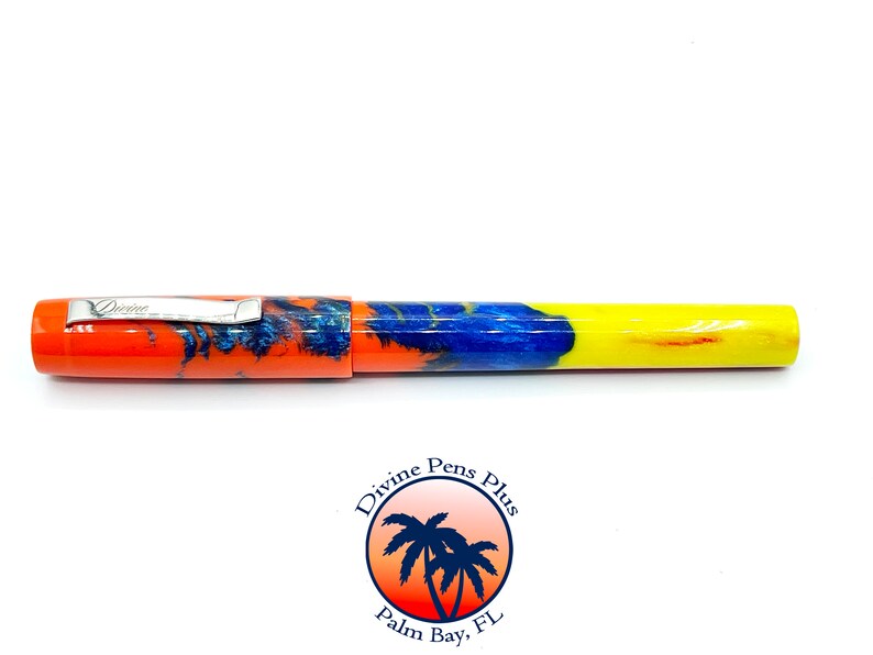 May include: A colorful pen with a vibrant design. The pen features an orange cap and barrel, transitioning to blue and yellow. The pen has a silver clip with the word "Divine" engraved on it. The pen is a writing instrument.