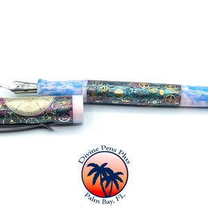 Puede incluir: Pluma estilogr&aacute;fica abierta con un dise&ntilde;o &uacute;nico. La pluma presenta un cuerpo azul y blanco veteado y un capuch&oacute;n con un dise&ntilde;o de esfera de reloj y engranajes. La pluma est&aacute; sobre un fondo blanco.