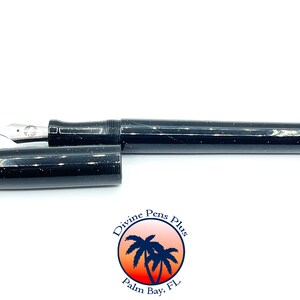 May include: A black fountain pen with white speckles, the cap is off, revealing the silver nib. The pen is lying horizontally on a white surface. The pen has a shiny finish. The logo for Divine Pens Plus is in the lower right corner.
