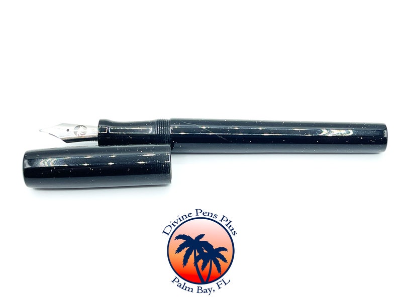 May include: A black fountain pen with white speckles, the cap is off, revealing the silver nib. The pen is lying horizontally on a white surface. The pen has a shiny finish. The logo for Divine Pens Plus is in the lower right corner.