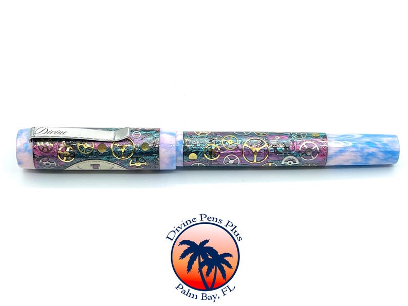 Puede incluir: Pluma detallada con dise&ntilde;o steampunk. La pluma presenta un clip plateado con la palabra "Divine" grabada. El cuerpo est&aacute; decorado con engranajes y una mezcla de colores, incluyendo morado, dorado y azul. El extremo de la pluma tiene un dise&ntilde;o veteado azul y blanco.