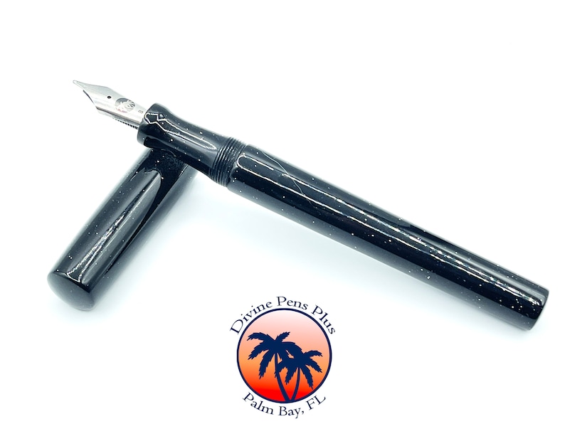 May include: A black fountain pen with white speckles and a silver nib. The pen's cap is open, revealing the nib. The pen is on a white background. The pen has the Divine Pens Plus logo.