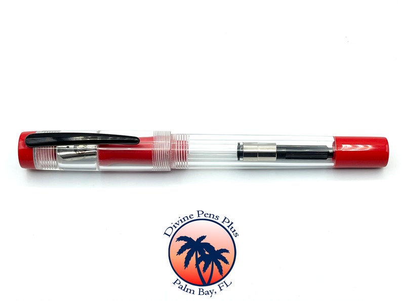 May include: A clear fountain pen with red accents. The pen has a transparent barrel, a black clip, and red ends. The nib is visible, and the pen appears to be new. The pen is a writing instrument.