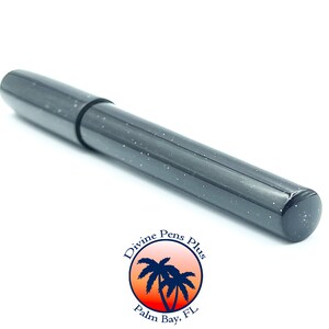 May include: A dark gray pen with white speckles, lying diagonally on a white surface. The pen has a cap and a cylindrical body. The pen is from Divine Pens Plus, Palm Bay, FL.