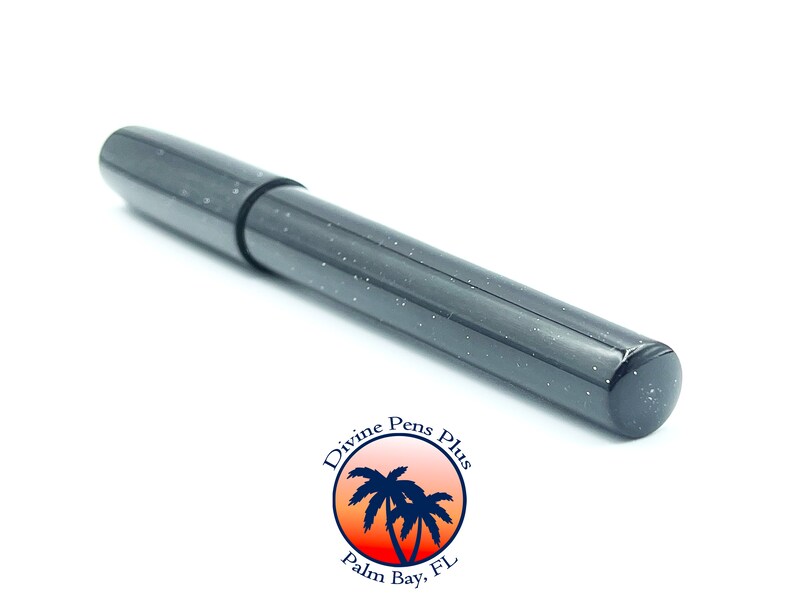 May include: A dark gray pen with white speckles, lying diagonally on a white surface. The pen has a cap and a cylindrical body. The pen is from Divine Pens Plus, Palm Bay, FL.
