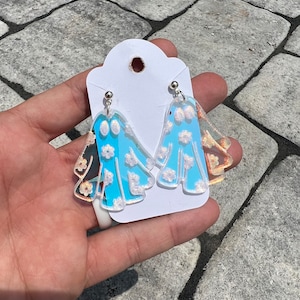 May include: A pair of ghost-shaped earrings. The earrings are light blue with white floral accents and a silver post. The earrings are held against a white card.