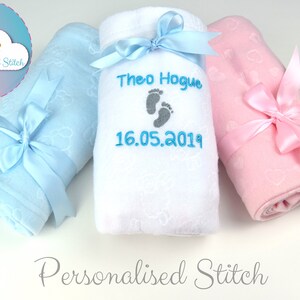May include: Three personalized baby blankets, one blue, one white, and one pink, with embroidered footprints and the name "Theo Hogue" and the date "16.05.2019".