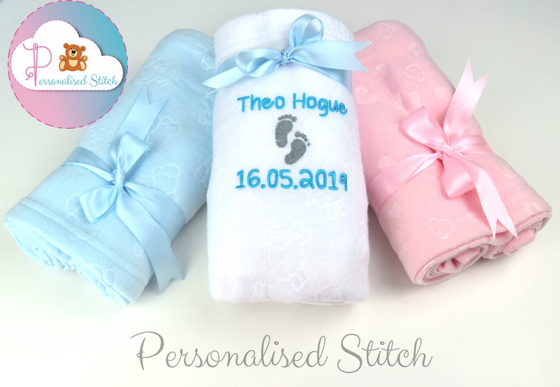 May include: Three personalized baby blankets, one blue, one white, and one pink, with embroidered footprints and the name "Theo Hogue" and the date "16.05.2019".