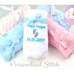 May include: Three baby blankets, one white, one light blue, and one pink, are rolled up and tied with ribbon. The white blanket has the name "Theo Hogue" and the date "16.05.2019" embroidered on it in blue thread.