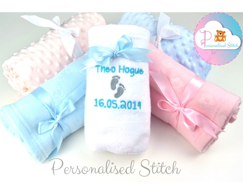 May include: Three baby blankets, one white, one light blue, and one pink, are rolled up and tied with ribbon. The white blanket has the name "Theo Hogue" and the date "16.05.2019" embroidered on it in blue thread.
