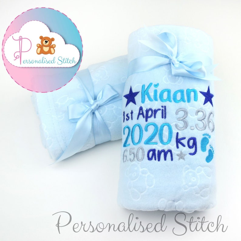 May include: A light blue baby blanket with a personalized embroidery design. The embroidery includes the name "Kiaan", the date "1st April 2020", the time "6:50 am", the weight "3.36 kg", and a pair of footprints.