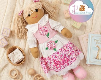 May include: A fabric doll with blonde yarn hair in pigtails, wearing a white collared shirt and a pink dress featuring floral appliques and the name Ava embroidered in pink thread. The doll has pink shoes and is surrounded by wooden blocks and books.