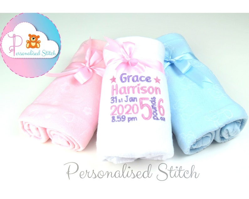 May include: Three personalized baby blankets, one pink, one white, and one blue, with a white ribbon tied around each. The white blanket has the text "Grace Harrison 31st Jan 2020 5 pounds 6 oz" embroidered on it.