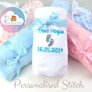 May include: A white baby blanket with blue ribbon tied around it. The blanket is embroidered with the name "Theo Hogue" and the date "16.05.2019". The blanket is surrounded by other baby blankets in pink and blue.