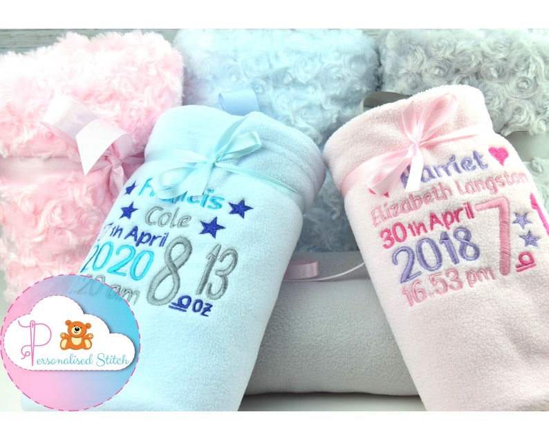 May include: Two personalized baby blankets, one blue and one pink, with embroidered names, birth dates, and times. The blankets are made of soft fleece and have a satin ribbon tied around them.