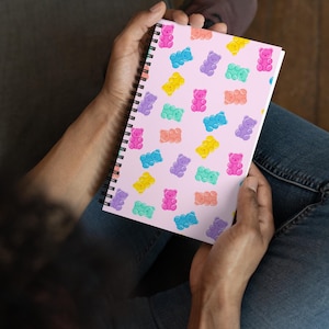 Puede incluir: Un cuaderno con espiral con una cubierta rosa claro que presenta un patrón de ilustraciones de ositos de goma coloridos. Los ositos de goma son en tonos de rosa, azul, amarillo, morado y naranja. El cuaderno está abierto, mostrando el diseño.