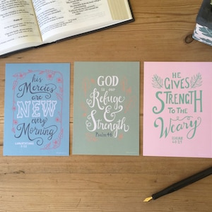 May include: Three inspirational cards with blue, green, and pink backgrounds. The cards feature hand-lettered text with bible verses. The first card says "His mercies are new every morning" with the verse "Lamentations 3:23". The second card says "God is our refuge and strength" with the verse "Psalm 46". The third card says "He gives strength to the weary" with the verse "Isaiah 40:29".