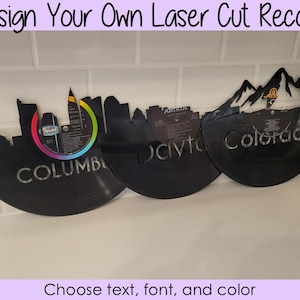 May include: Three laser cut vinyl records with city skylines and mountain ranges. The records are black and have white text. The text on the records reads "Columbia", "Dayton", and "Colorado".