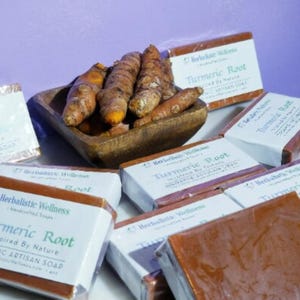 Puede incluir: Primer plano de un montón de raíz de cúrcuma, con varias barras de jabón artesanal de raíz de cúrcuma. El jabón está envuelto en papel blanco con el texto "Herbalistic Wellness" y "Turmeric Root" impreso en él.