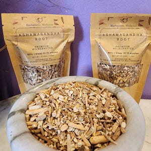 May include: Two brown paper bags filled with dried ashwagandha root. The bags have a white label with the text "Ashwagandha Root" and "Premium Wildcrafted". The text "85&deg;c | Steep 10-15 minutes" is also on the label. A white bowl filled with the dried root is in the foreground.