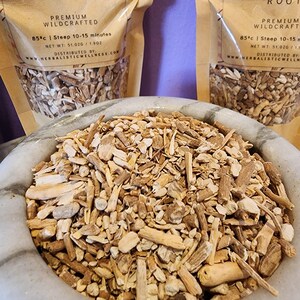 May include: A close-up of a bowl filled with dried, light brown, and beige root pieces. The bowl is made of white ceramic. The image is accompanied by two brown paper bags with text that reads "PREMIUM WILDCRAFTED 85&deg;c | Steep 10-15 minutes NET WT: 51.020 / 1.902 DISTRIBUTED BY www.HERBALISTICWELLNESS.COM".