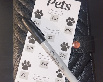 May include: A white card with the word "Pets" written in black at the top. The card has a grid of paw prints and bone icons with dollar amounts written next to them. The card also includes the text "Save$100" and "NorthStar Budgeting". A white dry-erase marker is lying on top of the card.