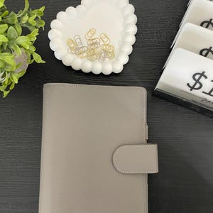May include: A light gray planner with a tab closure sits on a dark surface. A white heart-shaped dish with gold and silver paper clips is above the planner. A small plant is in the upper left corner.
