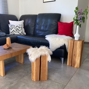 Puede incluir: Dos taburetes de madera con un cojín de piel sintética en la parte superior, colocados delante de un sofá de cuero negro con almohadas rojas y grises. Los taburetes están en una sala de estar con suelo de baldosas grises.