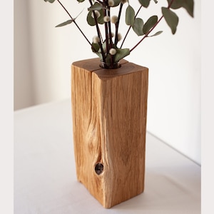 Könnte beinhalten: Eine rechteckige Holzblockvase aus hellbrauner Eiche mit sichtbarer natürlicher Holzmaserung und einem dunklen Astloch auf der Vorderseite. Ein kleiner Glaseinsatz ist oben mittig eingelassen und enthält mehrere Stiele getrockneten Eukalyptus.