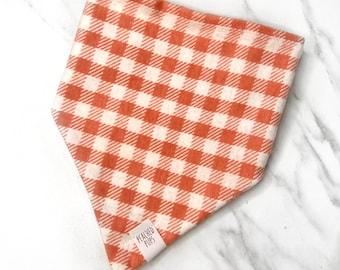 May include: Orange and white checkered dog bandana. The bandana is made of a soft fabric and features a classic gingham pattern. A small white tag with the text "PEACHED PUPS" is sewn on the corner.
