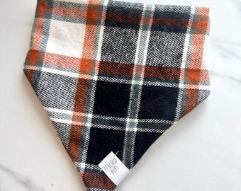 Op de afbeelding: Een driehoekige hondenbandana met een geruit patroon. Het patroon bevat oranje, zwarte, witte en grijze vierkanten. Een klein wit label met de tekst "PEACHED PUPS" is aan de bandana bevestigd.