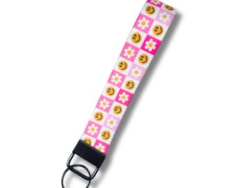 Pode incluir: Um porta-chaves de pulso com padrão rosa e branco com um anel de metal preto. O padrão apresenta quadrados alternados de rostos sorridentes e flores de margarida. A alça de pulso tem aproximadamente 15 cm de comprimento.