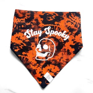 May include: Orange and black tie-dye dog bandana with the words "Stay Spooky" and a white skull graphic. The bandana is triangular in shape and has a small white tag with the words "Peached Pups".