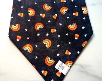 May include: A dark blue dog bandana featuring a repeating pattern of orange and yellow candy corn, orange and yellow rainbows, and white stars. A white tag with the text "PEACHED PUPS" is sewn on the bottom right corner.