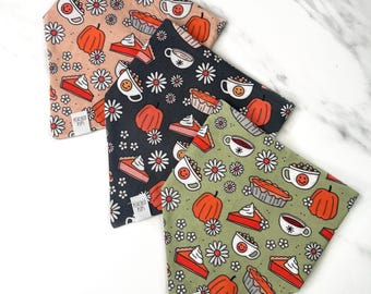Op de afbeelding: Drie hondenbandana's met herfstthema's. Elke bandana heeft een herhalend ontwerp van pompoenen, taartpunten, koffiekopjes met smiley-gezichten en madeliefjes. De bandana's zijn in tinten perzik, zwart en olijfgroen.