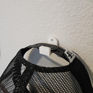 May include: A black mesh trucker hat suspended from a white wall-mounted hat holder. The hat features a black brim and a black mesh back. The hat holder has a simple, minimalist design.