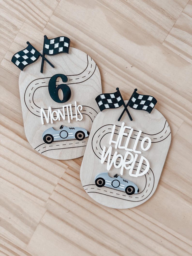 May include: Two wooden baby milestone signs featuring a race car theme. The first sign reads "6 Months" with a black number 6 and chequered flags. The second sign reads "Hello World" with chequered flags. Both signs have a black and white race track design and a grey race car.