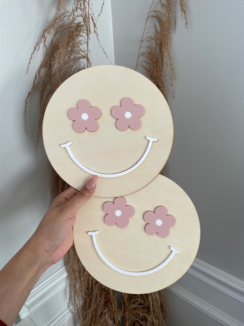 May include: Two wooden round wall decorations featuring a smiley face design. The eyes are pink flowers with white centres. The smile is white.