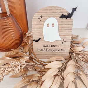 May include: A wooden Halloween countdown sign featuring a white ghost, black bats, and the text "days until halloween." The sign is surrounded by a gold-colored leaf wreath and sits near two orange glass vases.
