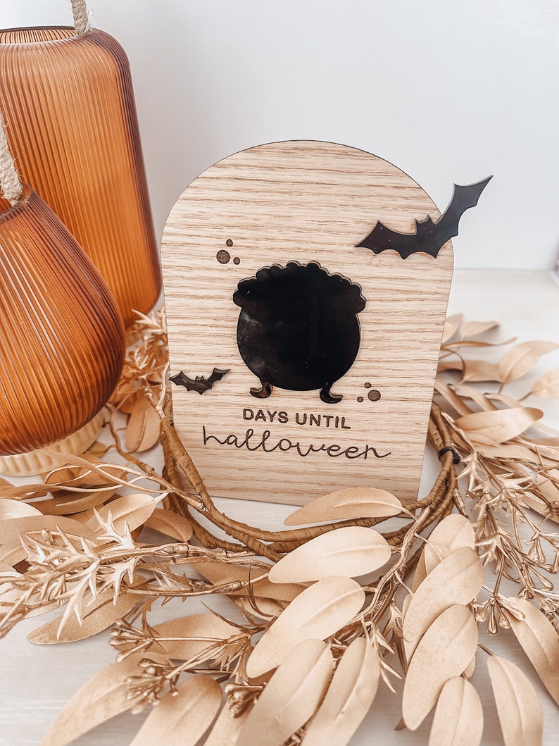 May include: Wooden Halloween countdown block with a black cauldron, bats, and the text "DAYS UNTIL halloween." The block is arched at the top and sits on a bed of gold-colored leaves and vines. Two orange glass vases are in the background.