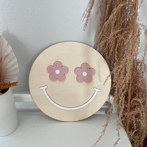May include: A wooden round sign with a white smiley face and pink flower eyes. The sign is on a white surface.