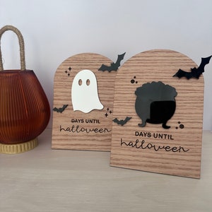 May include: Two wooden Halloween countdown blocks. One features a white ghost, the other a black cauldron. Both have black bat cutouts and the text "DAYS UNTIL halloween" in a script font. A brown glass jar with a rope handle is on the left.