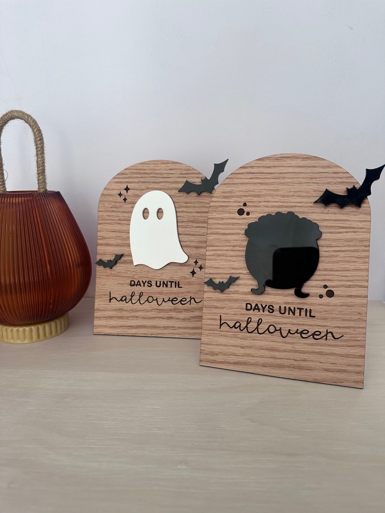 May include: Two wooden Halloween countdown blocks. One features a white ghost, the other a black cauldron. Both have black bat cutouts and the text "DAYS UNTIL halloween" in a script font. A brown glass jar with a rope handle is on the left.