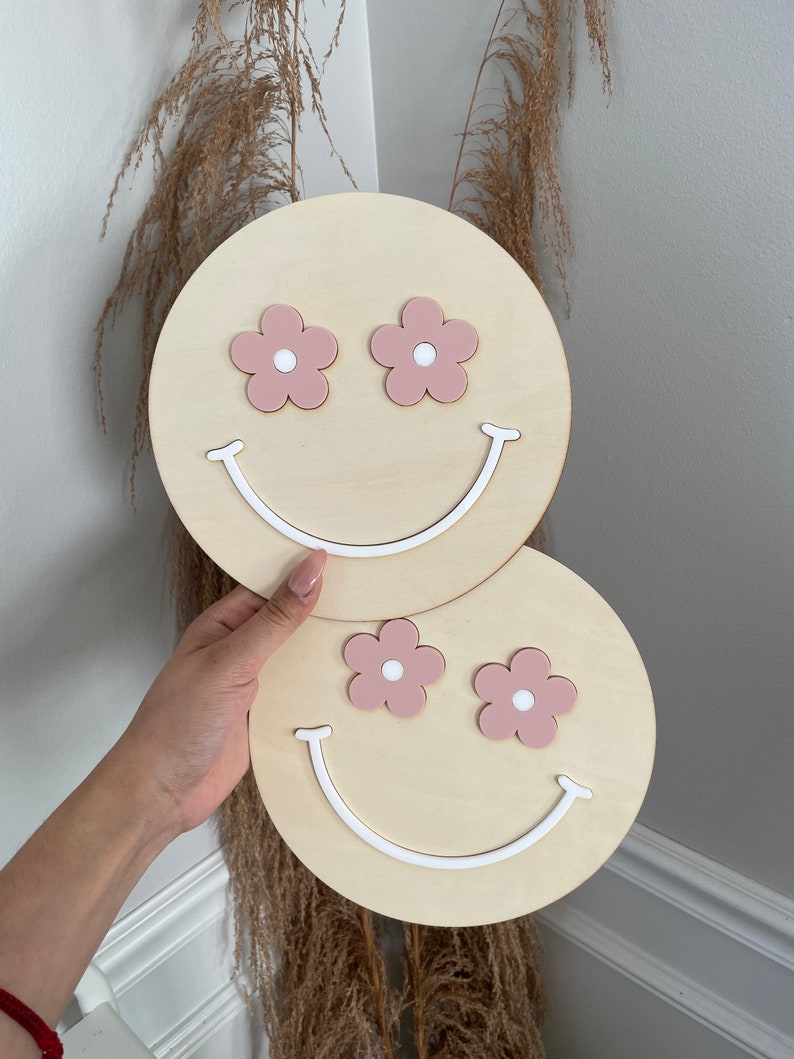 May include: Two wooden wall decorations shaped like smiley faces with pink flower eyes and white smiles.