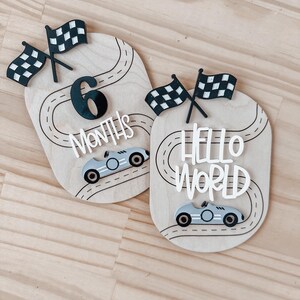 May include: Two wooden baby milestone signs featuring a race car theme. The first sign reads "6 Months" with a black number six and a black and white chequered flag. The second sign reads "Hello World" with a black and white chequered flag. Both signs have a black and white race track design and a silver race car.